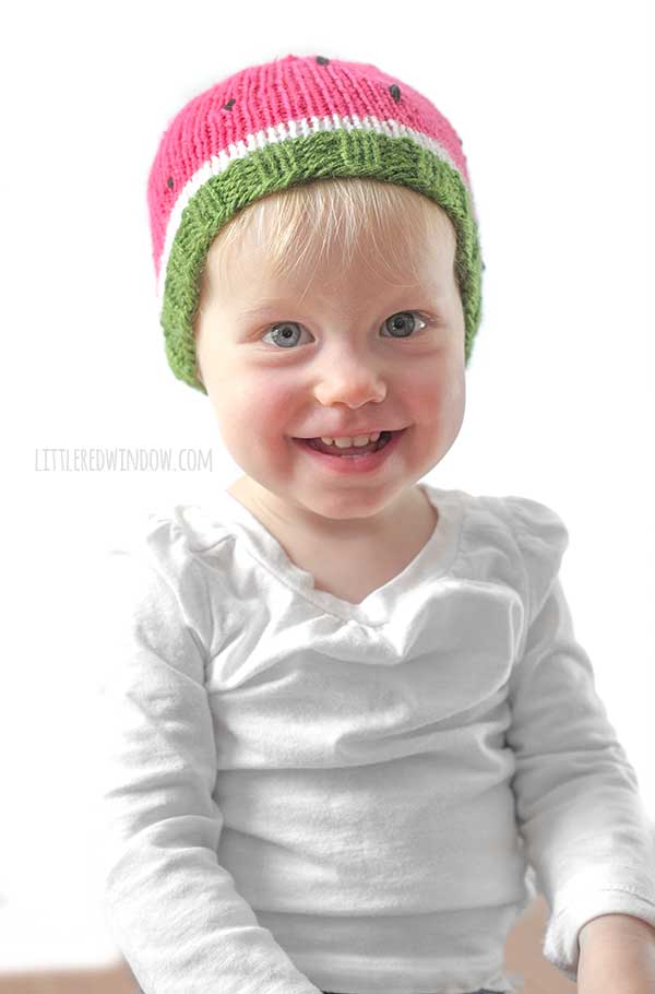 smiling baby in white shirt wearing a knit watermelon hat in green white and pink with black knit seeds in front of a white background