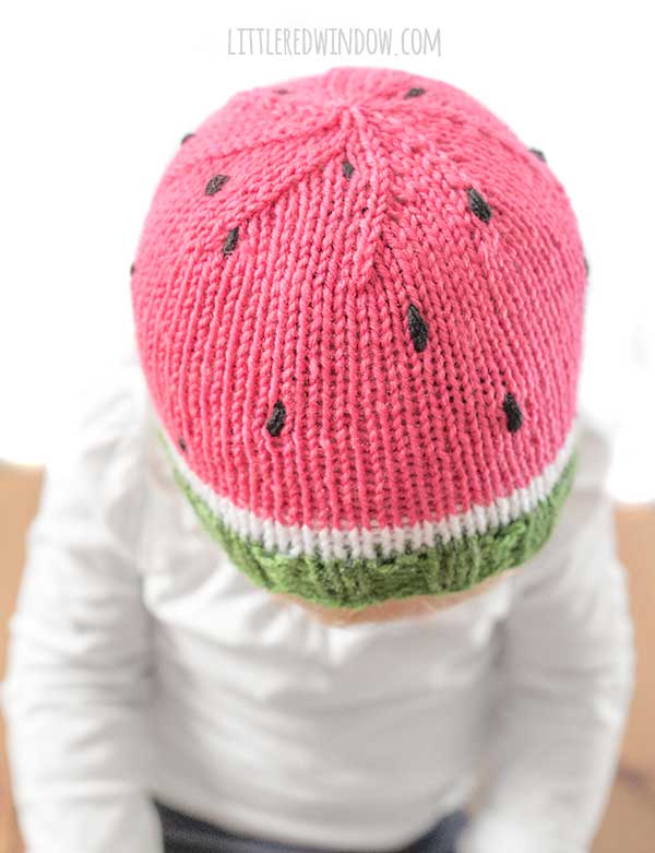 top view of child wearing a knit watermelon hat on a wood surface in front of a white background