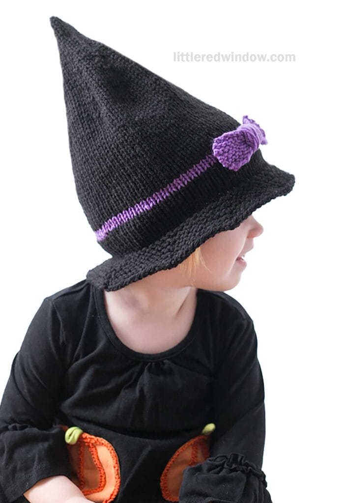 A child wearing a black, pointy witch hat adorned with a purple band and bow sits and turns to the side. Dressed in a black outfit with orange detailing, their light-colored hair stands out against the white background.