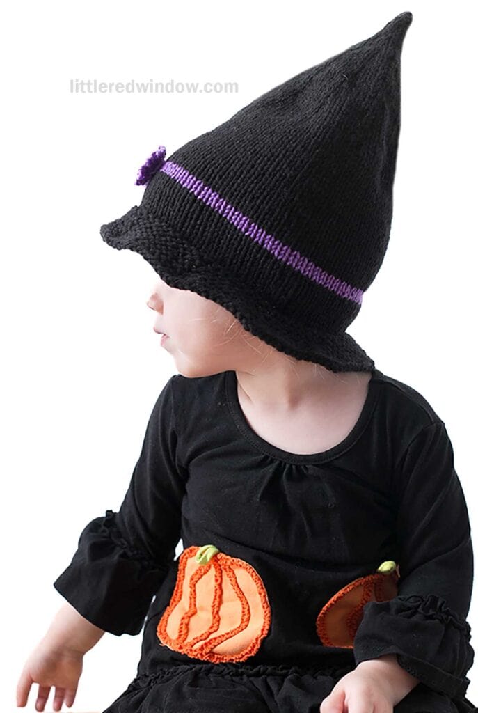 A young child wearing a black dress with orange pumpkin designs on the front is seen facing to the left. The child is also wearing a witch hat with a purple band, resembling those you might find in a knitting pattern. The background is plain white.