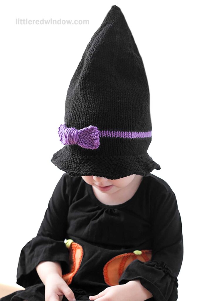 A young child wearing a black pointed witch hat with a purple bow, dressed in a black outfit with pumpkin designs on the front, sits against a white background. The child is looking down and smiling softly, as if dreaming about a new witch hat.