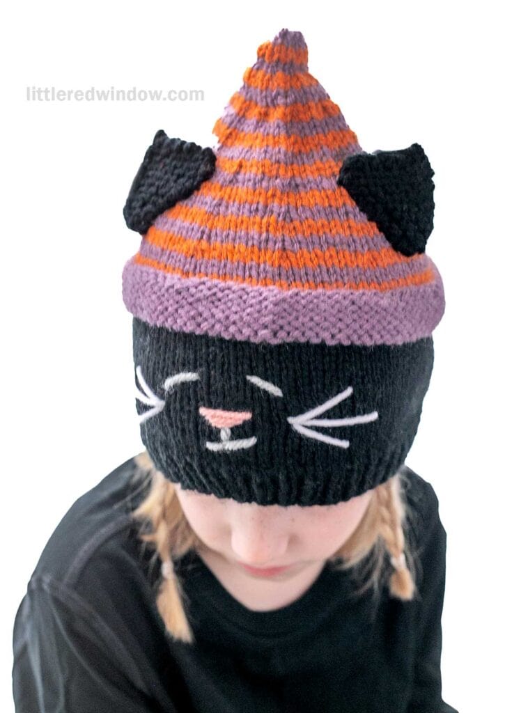 A person is wearing a black, purple, and orange knitted hat designed to look like a cat. The hat features black cat ears, embroidered whiskers, and a triangular orange and purple striped top. The person is also wearing a black shirt.