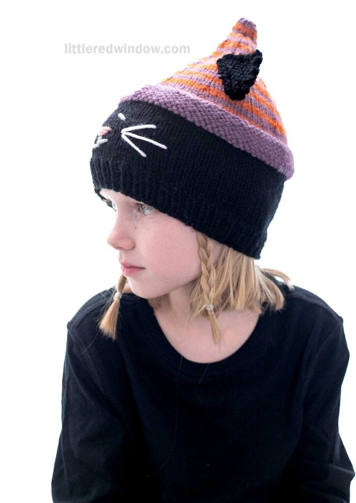 A child with blonde hair and braids is wearing a knitted hat designed to look like a cat, featuring orange and black stripes, cat ears, and embroidered whiskers. They are dressed in a black top, looking to the side against a white background.