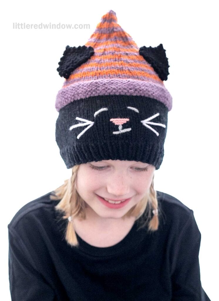 A young girl with blonde hair is smiling while wearing a knitted hat designed to look like a cat. The hat has black cat ears, a striped pattern in orange and purple, and a black lower portion with a cat face featuring whiskers and a pink nose.