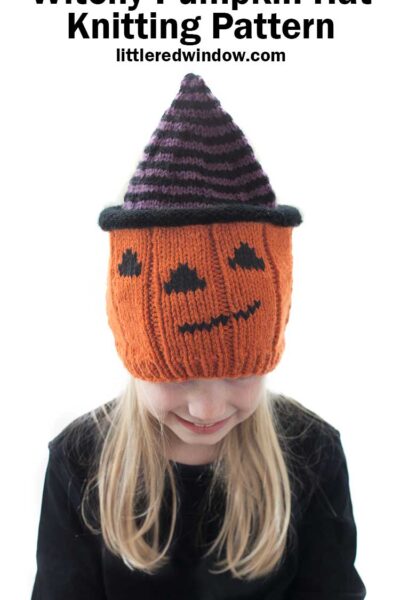 Smiling blond girl wearing a knit hat that looks like a jack o lantern wearing a purple and black striped witch hat while she is is looking down