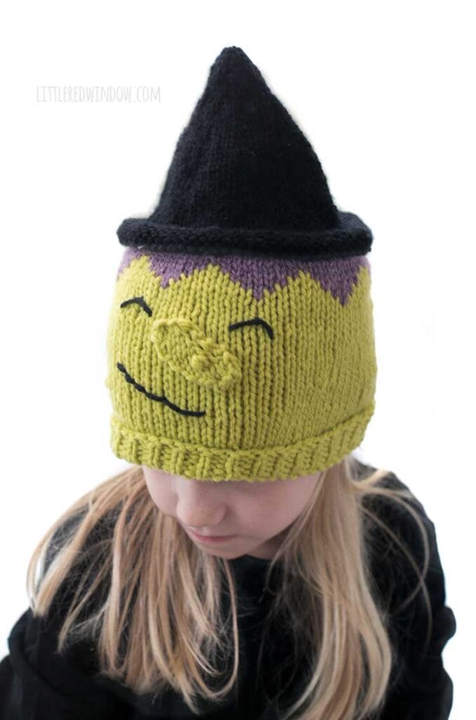 Little blond girl in a black shirt wearing a hat that looks like a witch including green witchy skin warty nose purple hair and black pointy witch hat while the girl is looking down and to the left