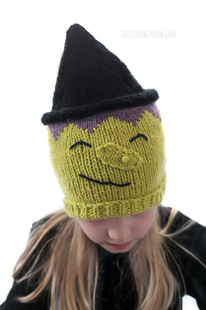 Closeup of Little blond girl in a black shirt wearing a hat that looks like a witch including green witchy skin warty nose purple hair and black pointy witch hat while the girl is looking down