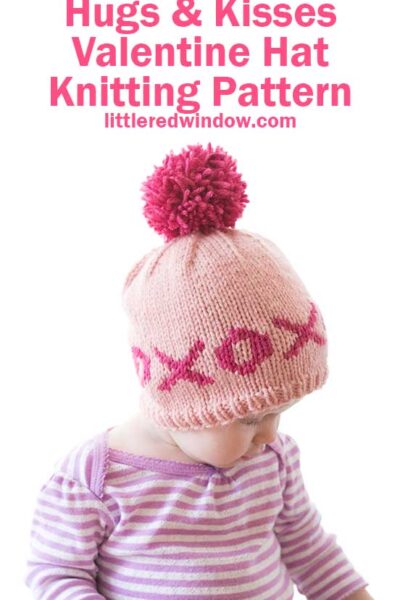 A baby in a striped shirt wears a pink knitted Valentine hat adorned with a red pom-pom and "XOXO" pattern, set against a white background. The text "Hugs & Kisses Valentine Hat Knitting Pattern" and "littleredwindow.com" gracefully floats above.